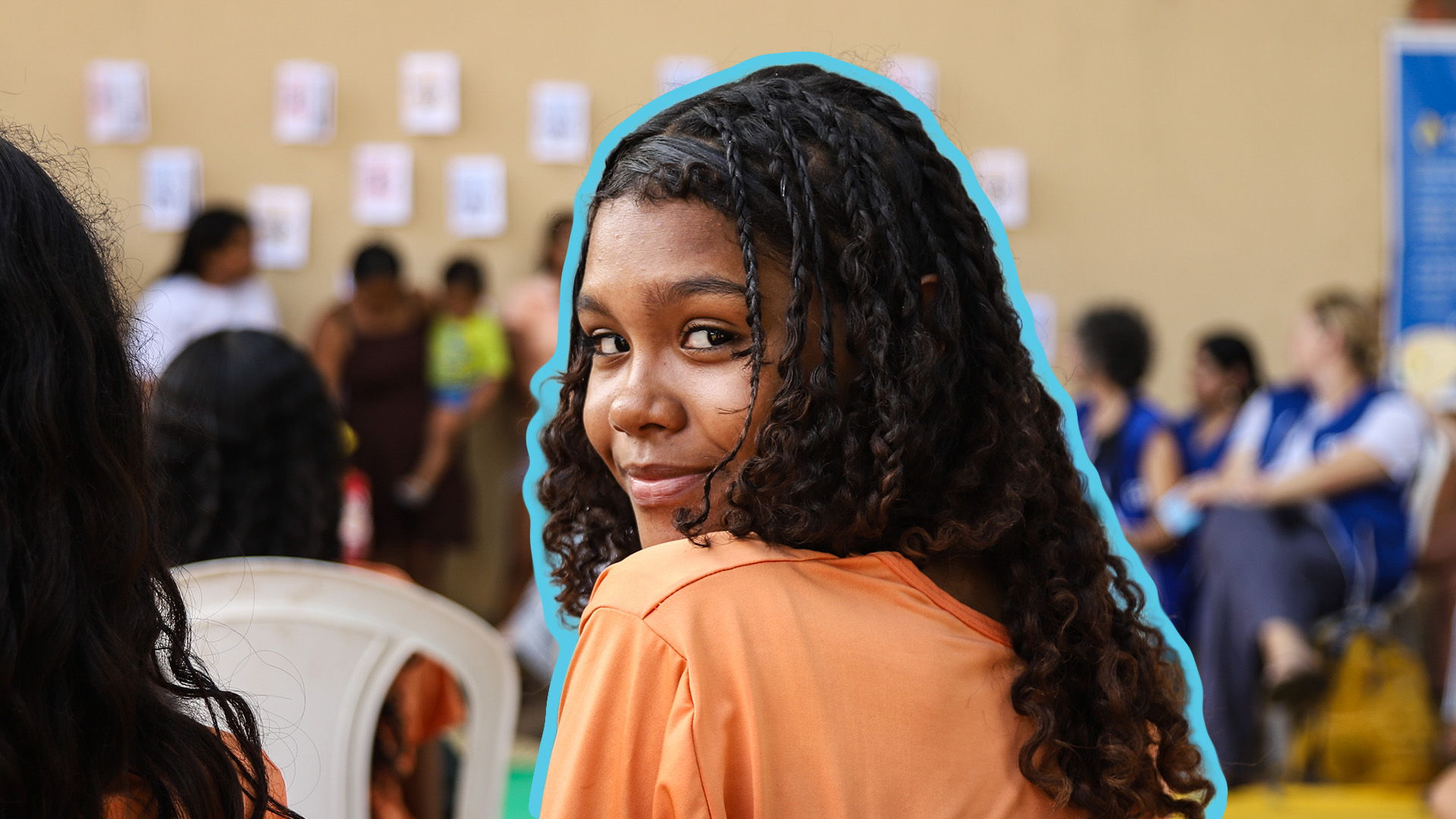 A smiling young girl with long curly hair wearing an orange top.