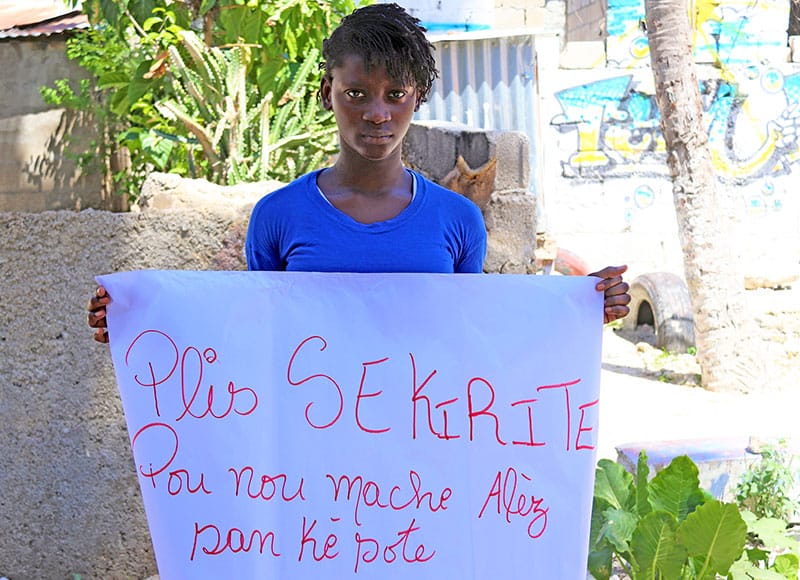 Chrisderlie, a girl in Haiti, holds a sign written in Haitian Creole asking for "more security so we can walk without anxiety" (Plus sekirite pou nou mache alez san ke sote)