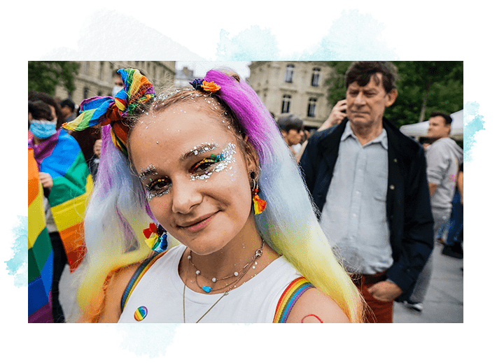 A young person with long hair dyed the colors of the rainbow smiles for the camera at what looks like an LGBTQ+ celebration. Plan International USA partnering with Unsplash.