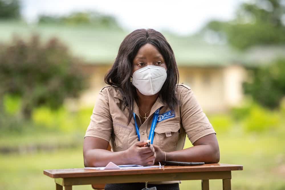 A woman in Zimbabwe sits at a desk outside, wearing brown shirt and face mask.