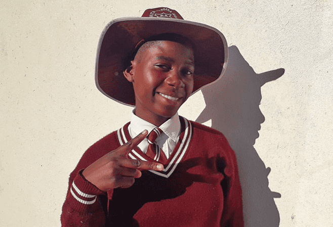 A girl in Zimbabwe with a hat and red sweater makes a peace sign and smiles at the camera.