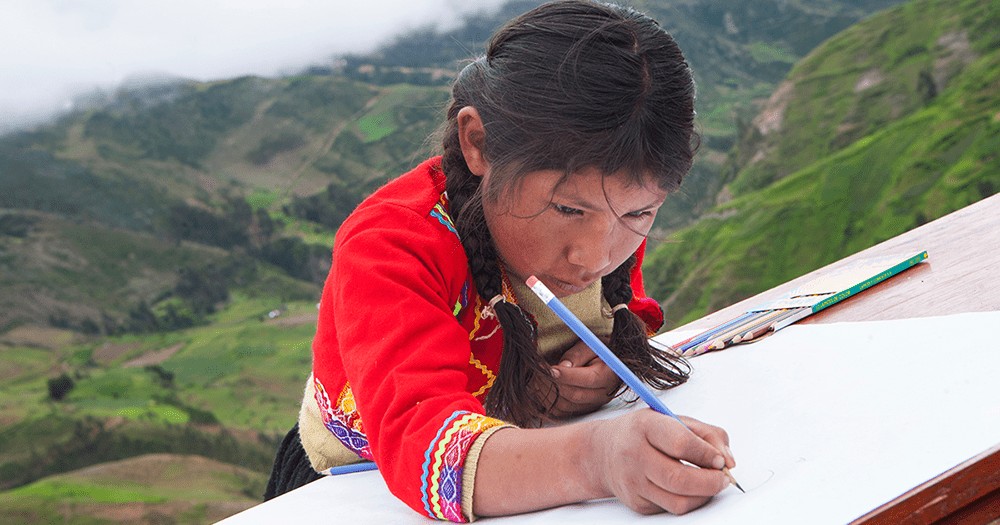 Girl wearing red embroidered top draws on large paper with a box of coloring pencils.