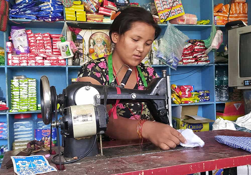 A young woman from Nepal sits at a sewing machine working on a white piece of cloth, with blue shelves of products to sell behind her.
