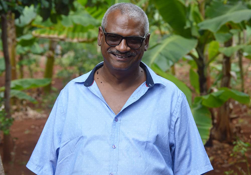 Raúl, a 55-year-old man from the Dominican Republic, smiles at the camera wearing black-rimmed glasses and a short-sleeved blue button-up shirt.