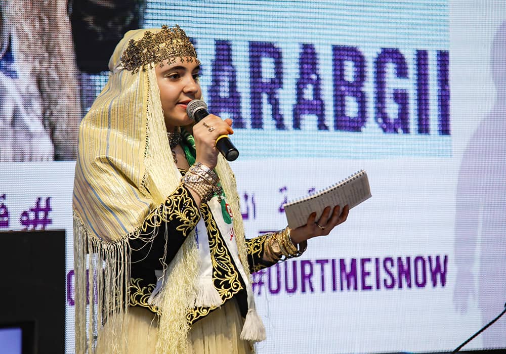 A girl wearing a light yellow head covering speaks into a microphone in her right hand while holding a small notebook in her right hand.