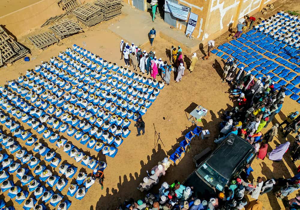 Aerial image of 1,800 blue food kits laid out on the dusty ground, surrounded by a crowd of people waiting to receive them.