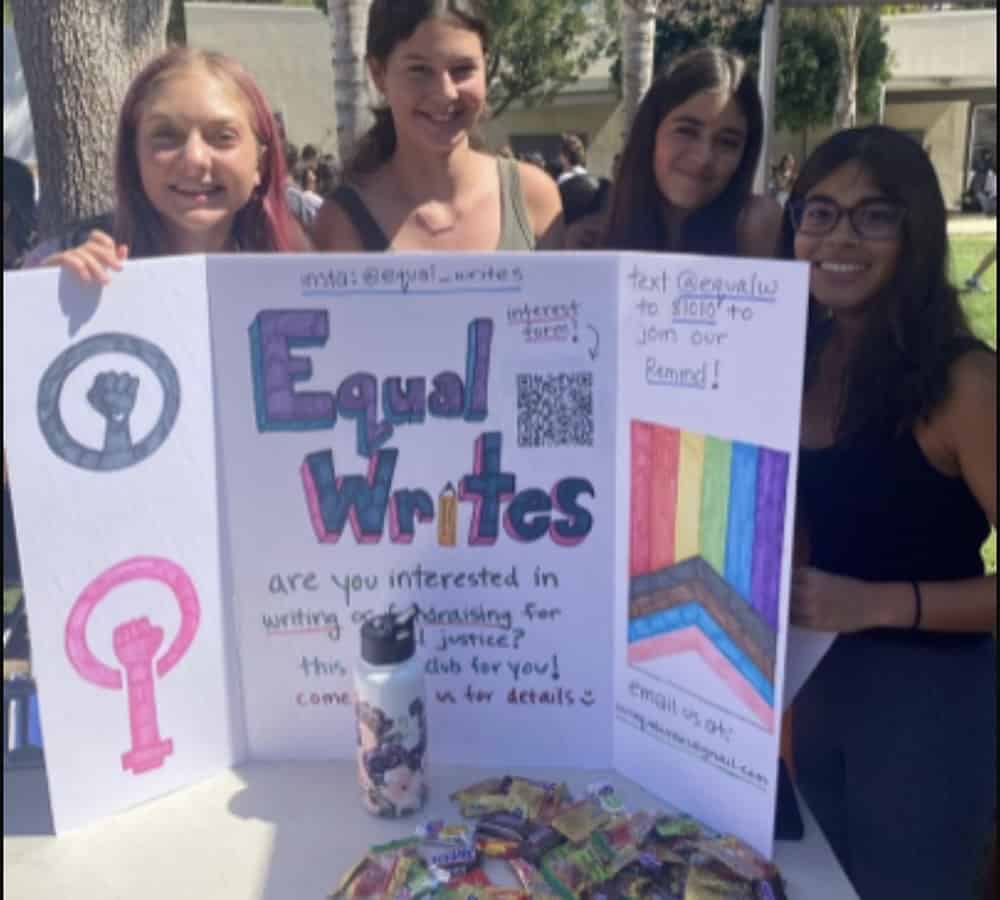 Four girls stand behind a white trifold project board with “Equal Writes” written in big block letters, next to a drawing of an inclusive rainbow pride flag.