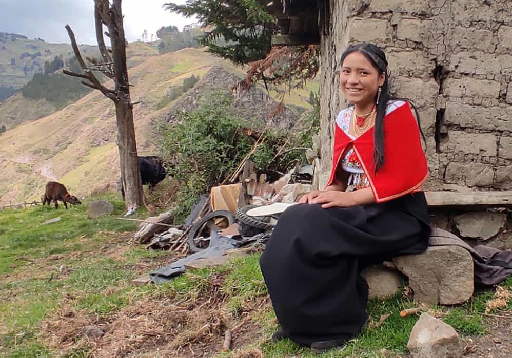 Mirella, a 16-year-old girl from Ecuador, sits on a rock outside, wearing a long black skirt and a traditional white Ecuadorian blouse covered by a red shawl.