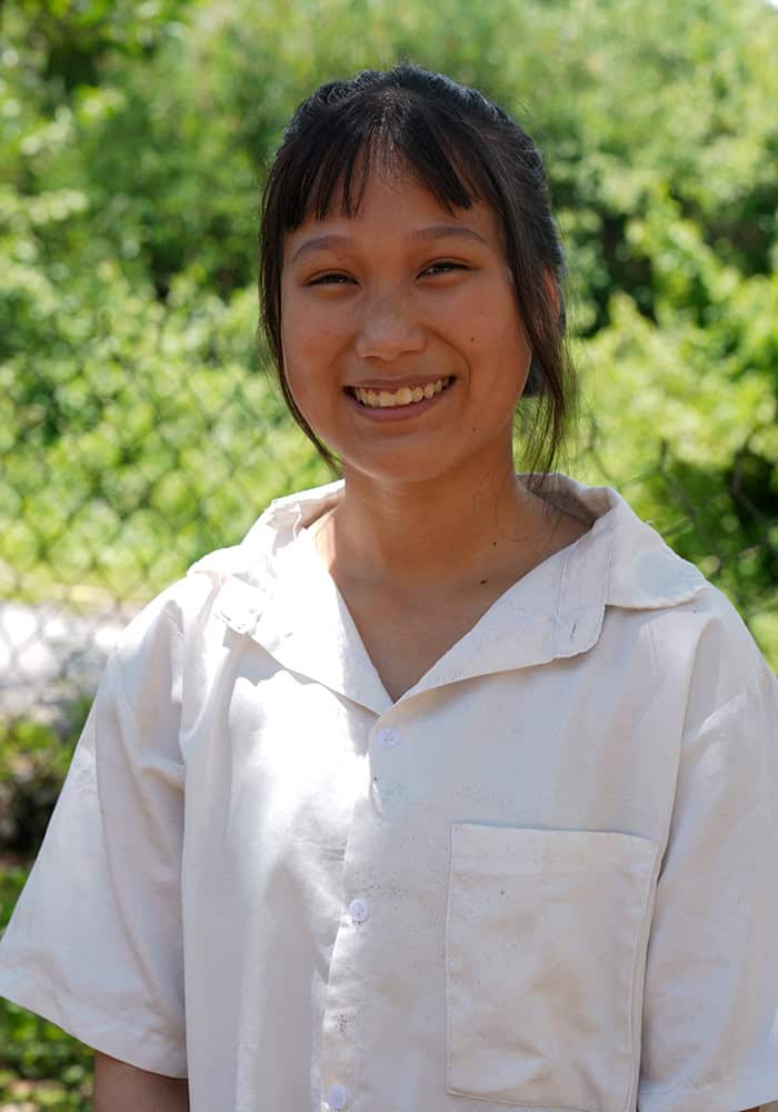 Nga, a 15-year-old Vietnamese girl, smiles as she stands in front of a fence in a white collared shirt.
