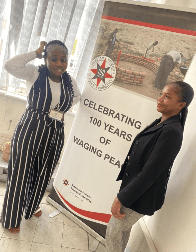 Tabeth and Michelle, panel members from Zimbabwe, stand next to a peace promotion poster at the pre-consultation meeting. 