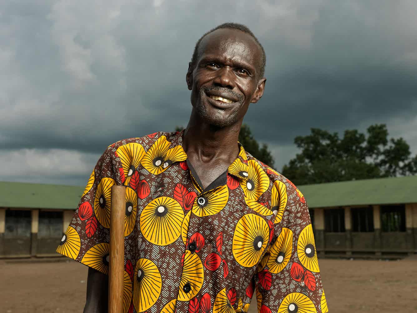 Clement, wearing yellow patterned shirt, holds a walking stick as he poses for a picture standing in front of the low green building where he teaches.
