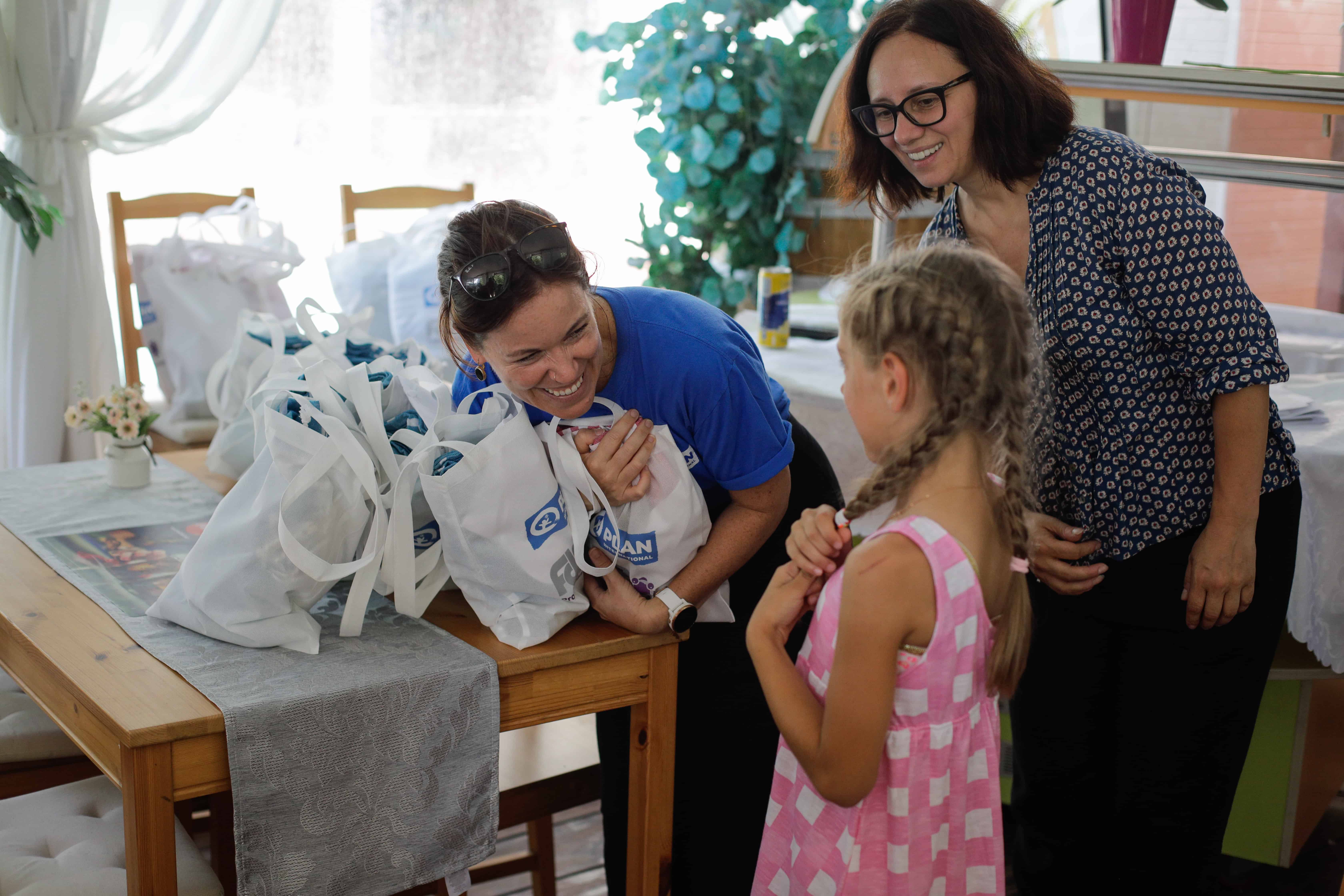 Colleen is leaning over a wooden table holding one of 10 or so white canvas bags with the Plan logo on them. She is smiling at a little girl, who is wearing a pink and white dress and has her dark blonde hair French braided.