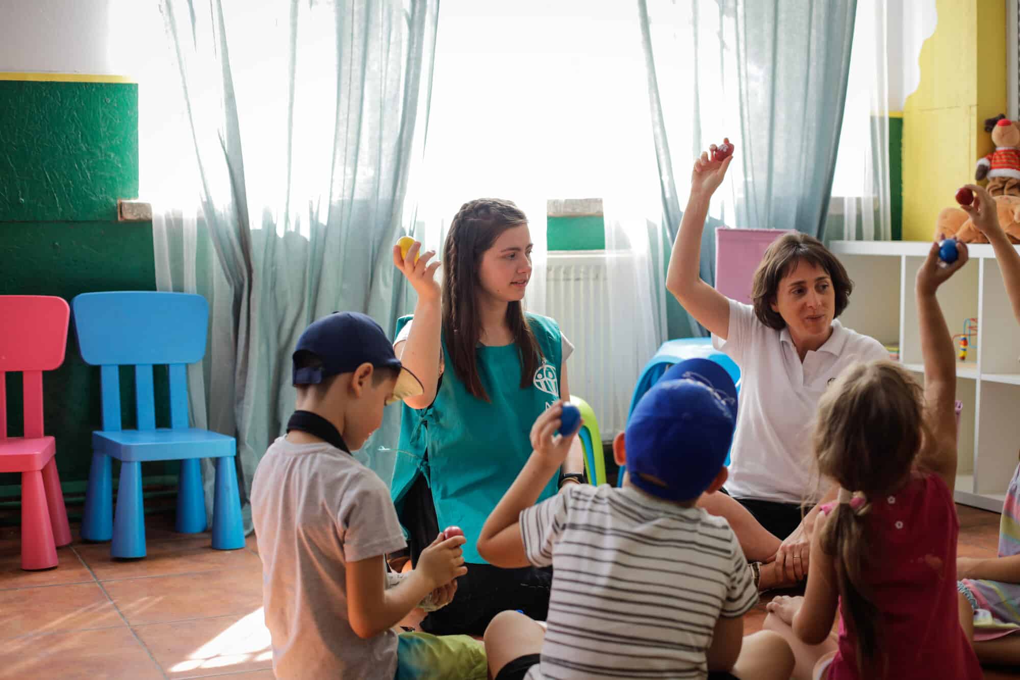 Cristina sits cross-legged on the floor wearing a white collared short-sleeved shirt and holding up a small red instrument high over her head in one hand. Five children sit on the floor in a circle around her, each with their own small musical instrument. 