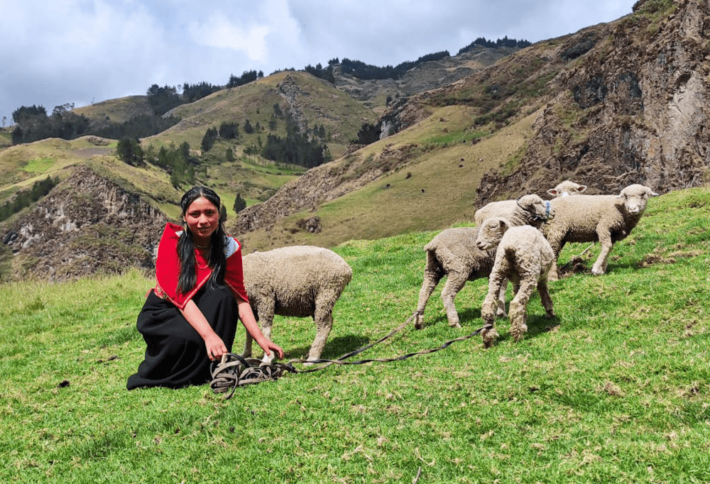 Mirella, a 16-year-old girl with her long black hair in two ponytails, tends to her family’s sheep in the mountains of Ecuador, wearing a red shawl and a long black dress.
