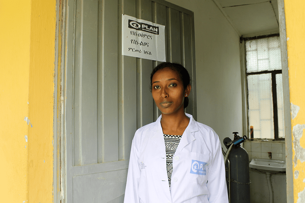 Bemenet, a young woman from Ethiopia wearing a white medical coat with Plan’s logo, stands in the doorway to a room where she provides counselling.