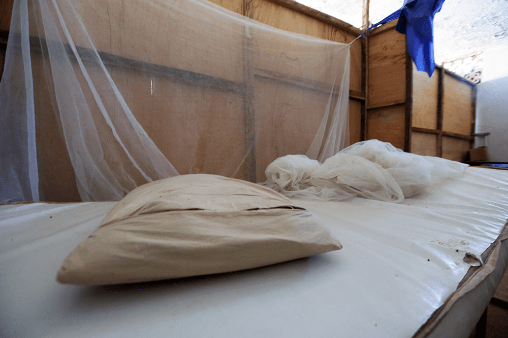 A simple white mattress cushion and pillow are used as a makeshift maternity bed, framed on one side by a short wooden partition that is partially covered with a white mosquito net. 