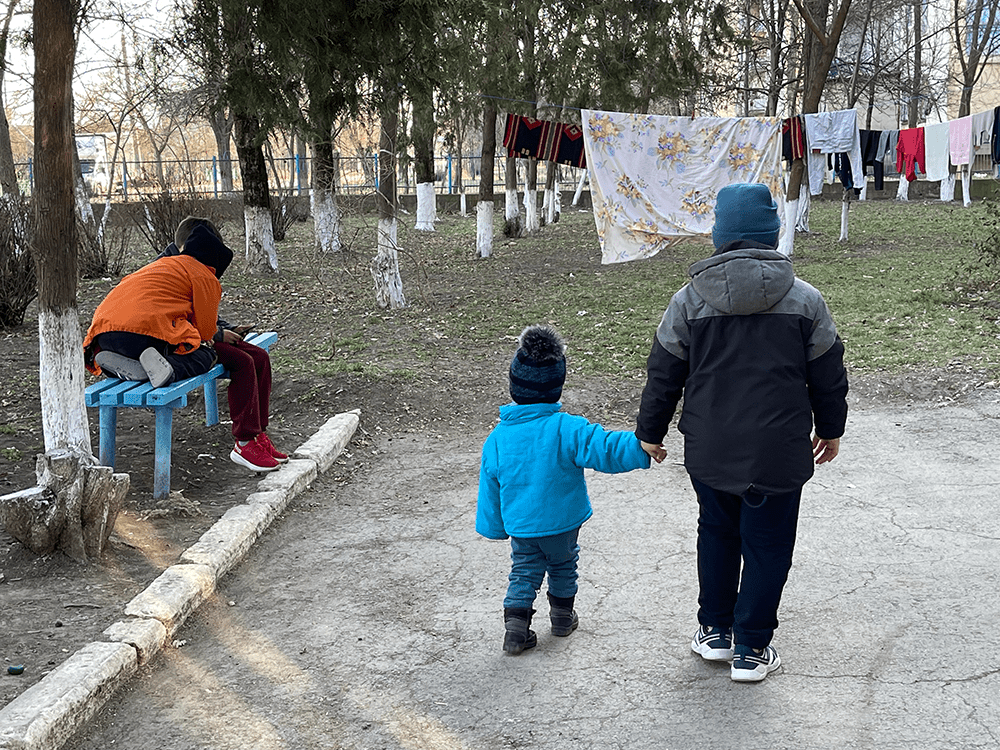 Two children wearing coats and knit hats walk together outside holding hands.