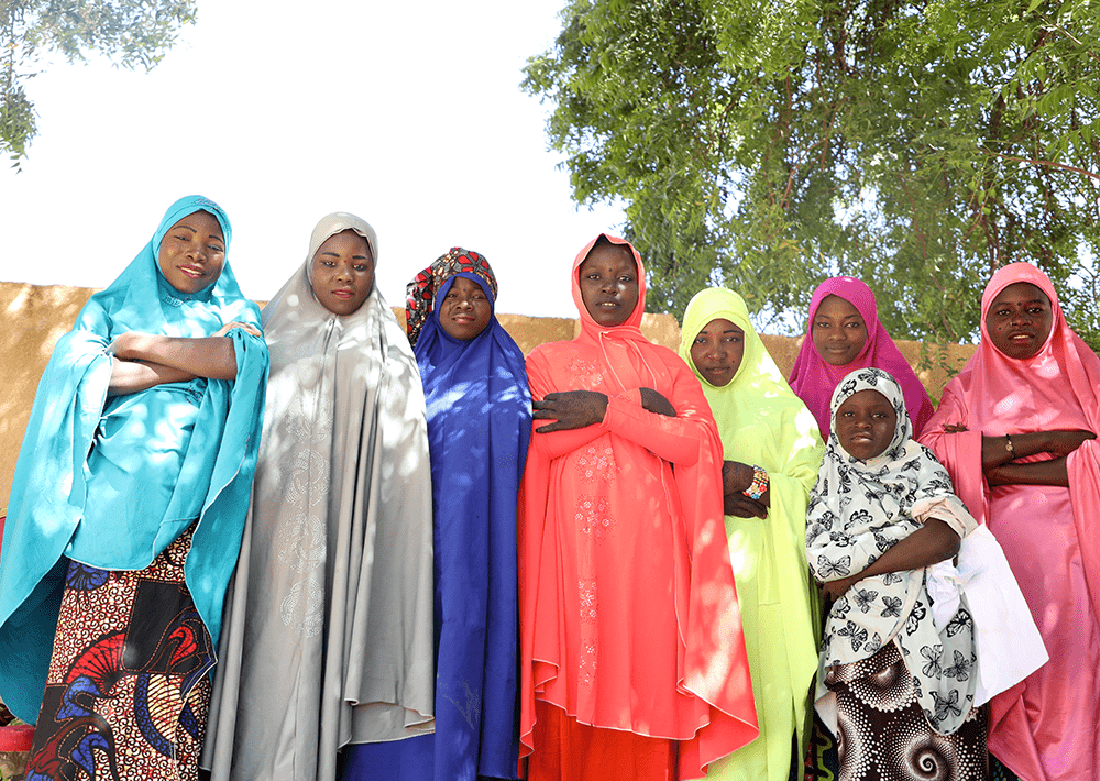 Eight girls from Niger wearing colorful shawls and headscarves stand together with their arms crossed. 