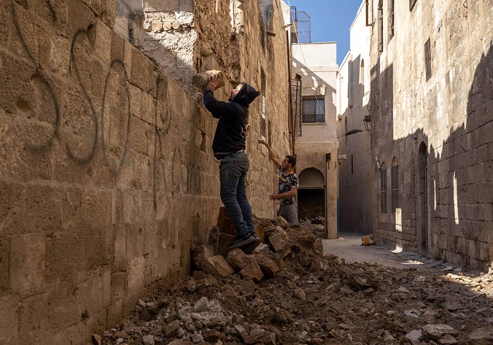 A man stands on building rubble wearing a black hooded sweatshirt and dark jeans, while holding a brick-sized piece of the building that has fallen.      
