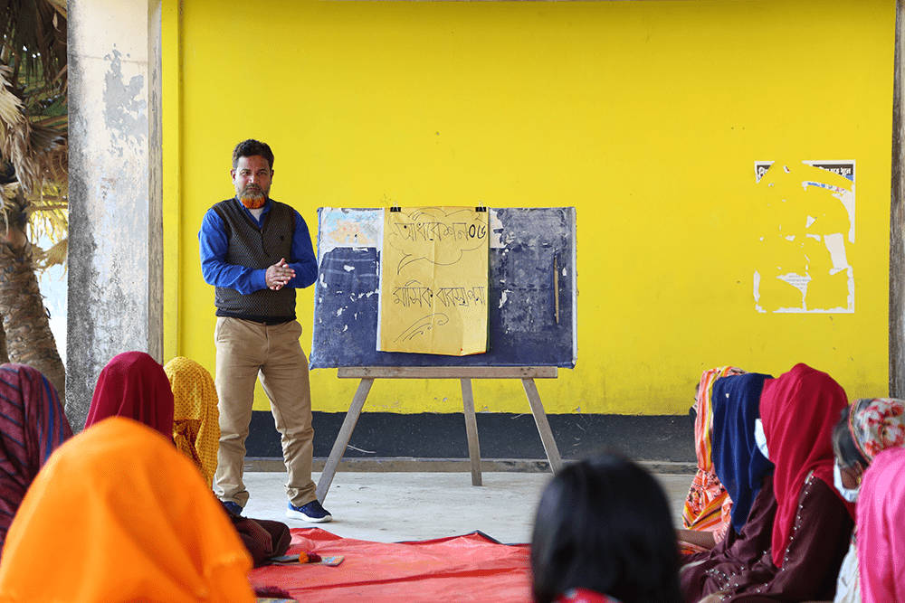 Abdul gives a lecture to girls in his class in front of a small, rectangular chalkboard perched on an easel with a large piece of paper clipped to it. 