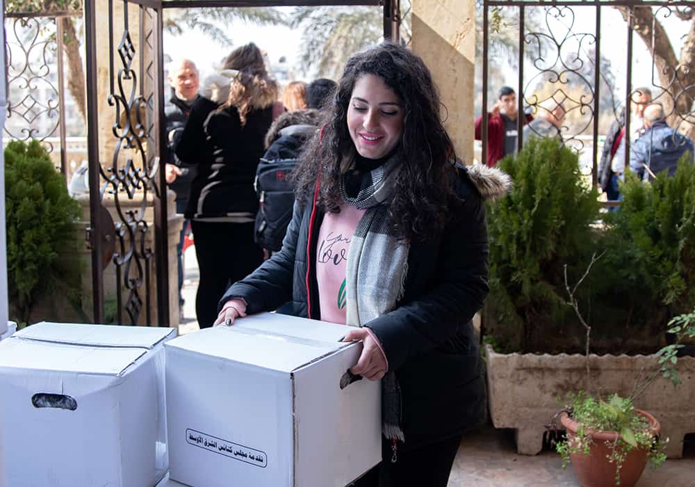 A woman wearing a pink sweatshirt, black and white checkered scarf and black zip-up jacket smiles as she picks up a white box, which is her hygiene kit. 