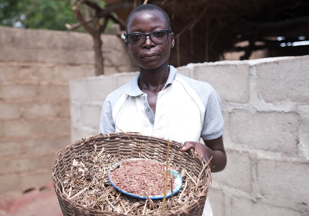Valentine wears glasses and a white shirt and holds a basket of animal feed. 