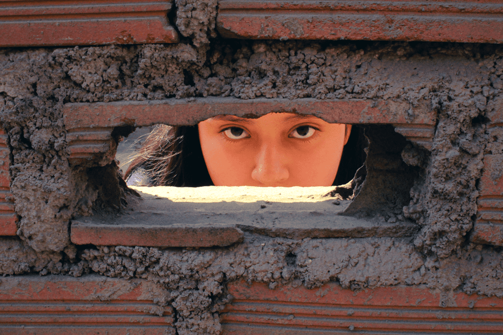 A girl’s eyes and nose are visible looking through a rectangular hole in a brick wall, looking directly at the camera.