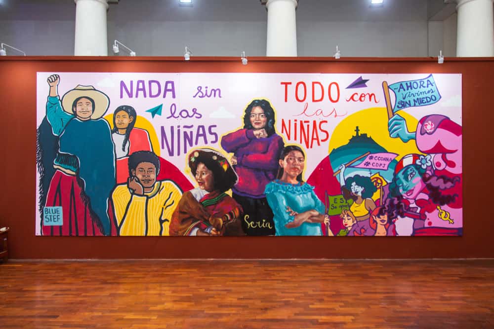 A colorful mural on a brown wall with the message: “Nothing without girls; everything with girls” in Spanish. 