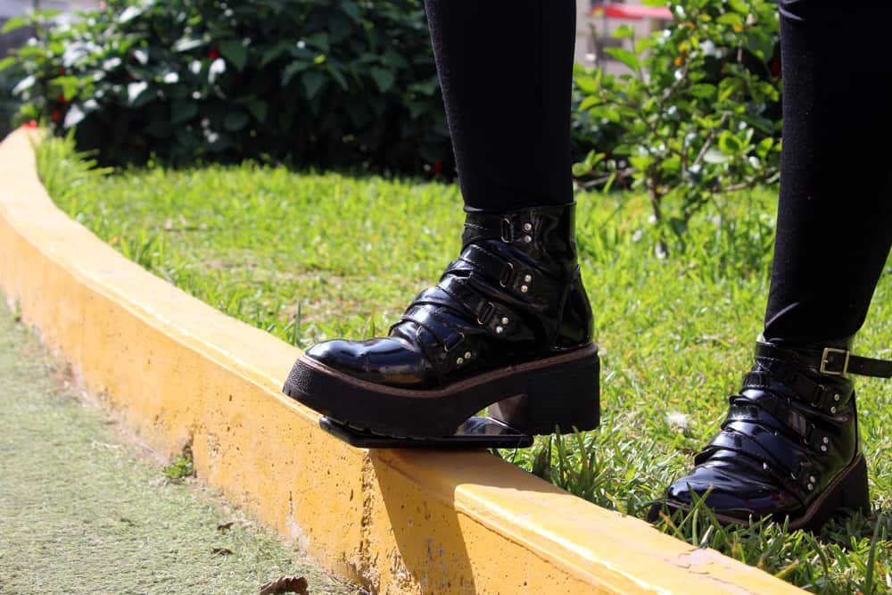 A girl wearing black tights and boots steps on a cell phone, which is perched on a yellow curb.