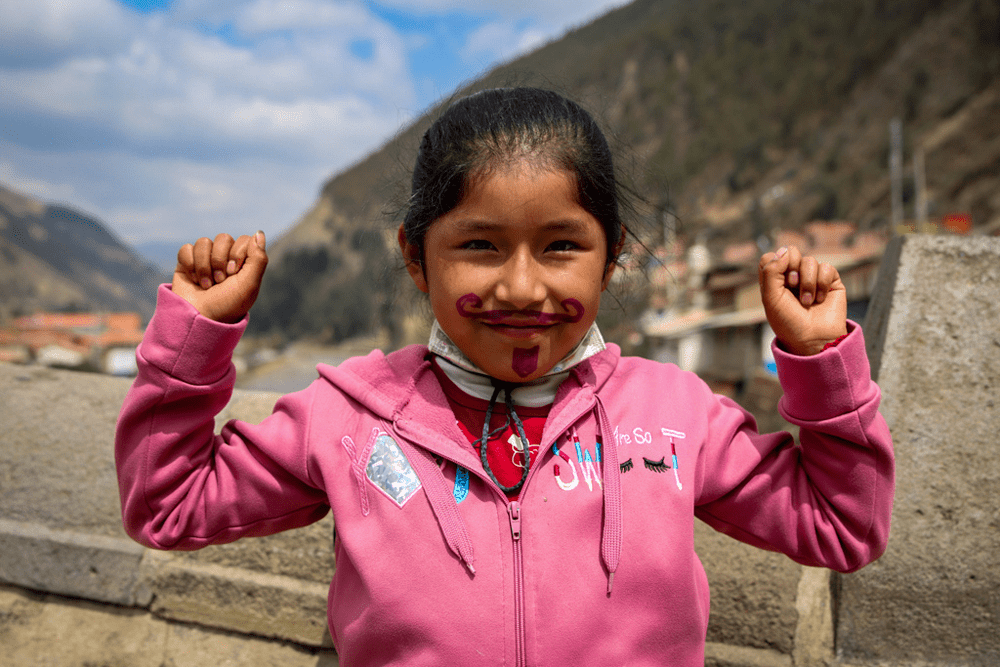 A girl wearing a pink sweatshirt with a fake mustache and goatee painted on her face holds up her arms and smiles at the camera.