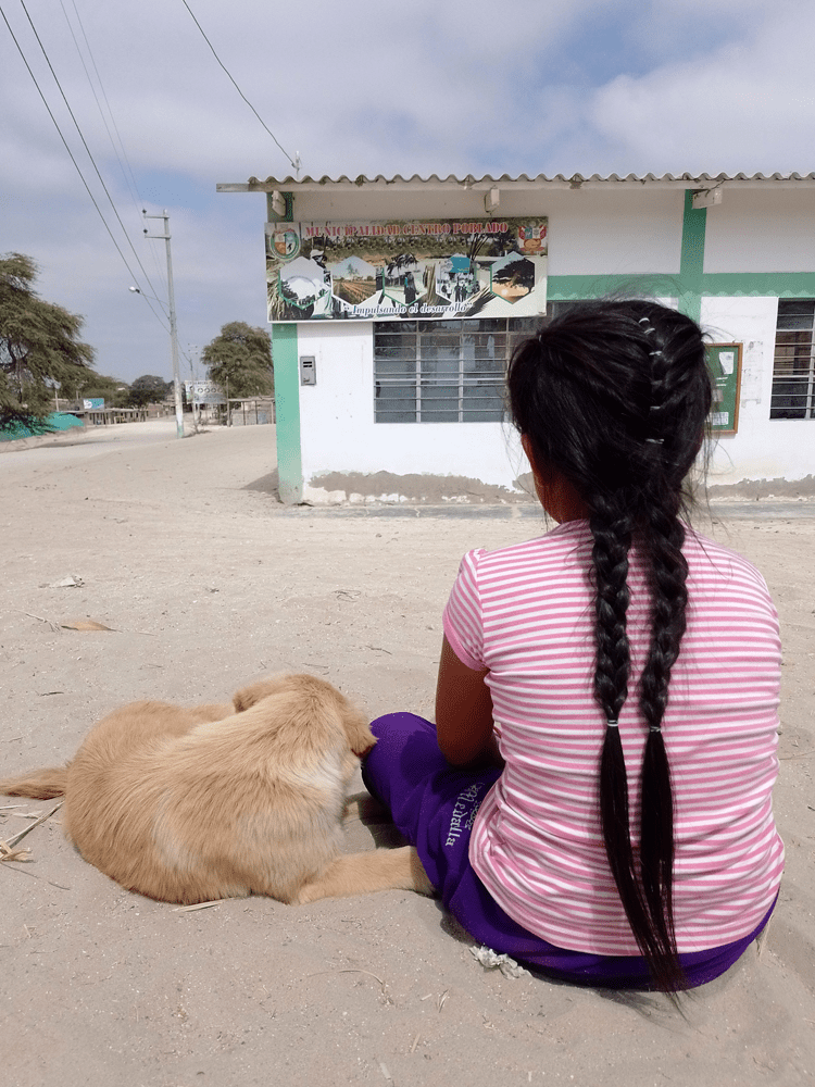 A girl with long hair in two braids and a pink striped shirt sits on the ground next to a light brown dog.