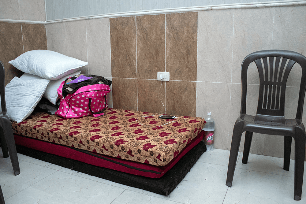 A mattress with a pile of pillows and a pink and white polka-dot bag placed in the corner of a tiled room with brown plastic chairs at the foot of and beside the makeshift bed.