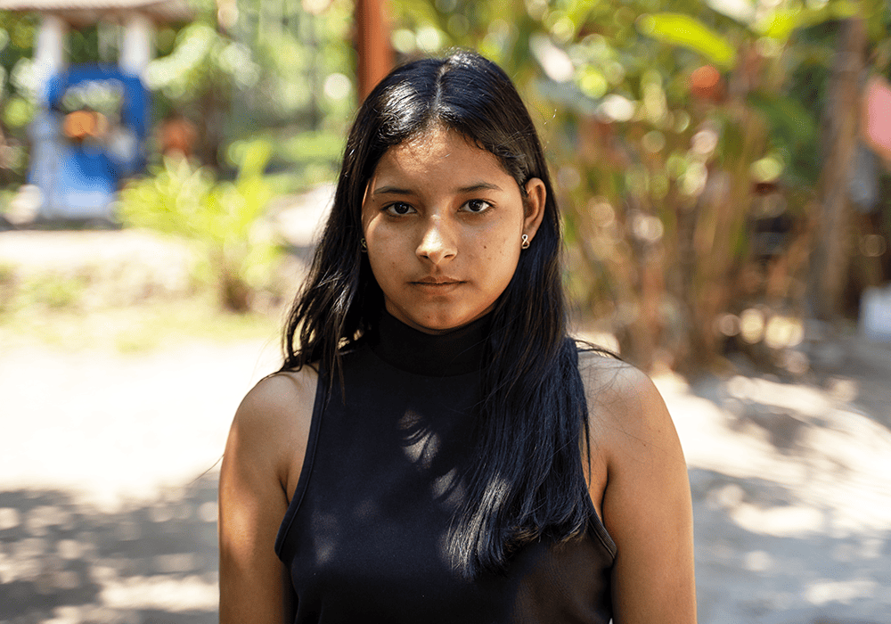 Carmen stands outside under the shade of a tree.