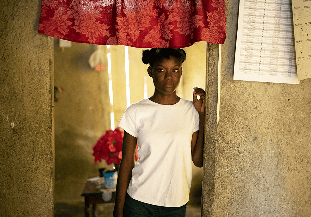 Dialissa stands in a doorway, with a red floral curtain hanging above her and papers pinned on the wall to her right. 