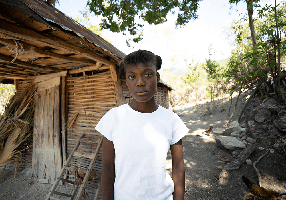 Dialissa wears a white tee-shirt and stands in front of a wooded shed.  