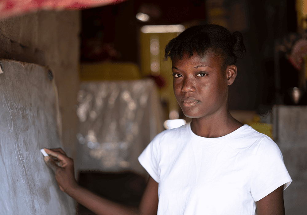 Dialissa looks at the camera while writing on a blackboard with chalk, in her house in Haiti. 