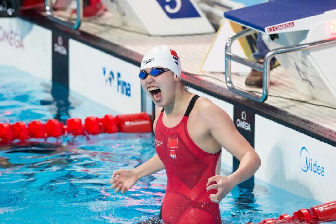 Fu Yuanhui, wearing a white swim cap, goggles and a red one-piece bathing suit representing China, stands in a pool between two floating red lane lines and screams in celebration.