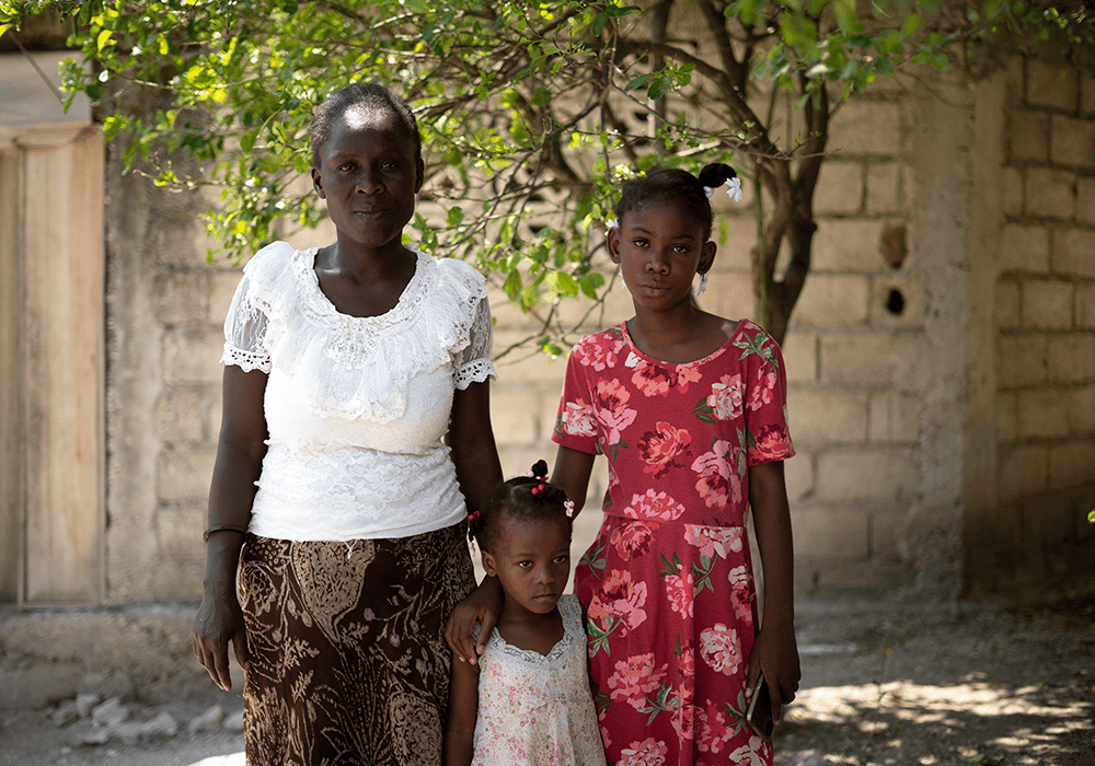 Lettycia stands under a tree next to her young cousin, who wears a pink and white dress, and her aunt stands to the left of them, who’s wearing a white shirt and brown skirt.