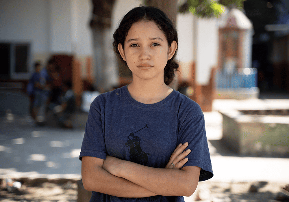 Miriam wears a blue t-shirt and stands outside with her arms crossed as she looks at the camera.