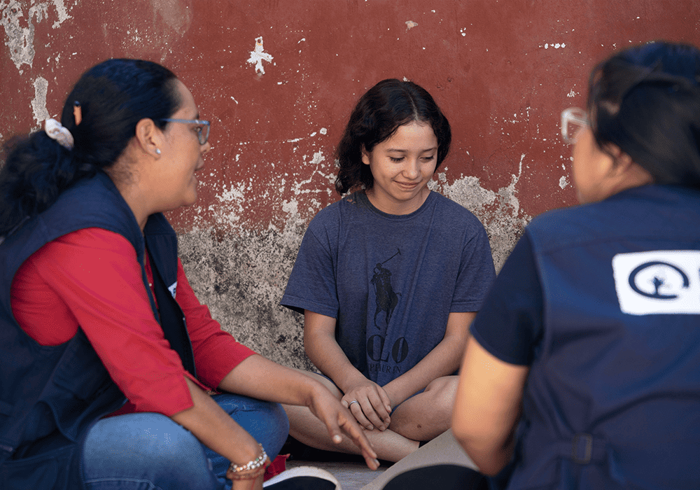 Miriam sits cross-legged on the ground in front of a red concrete wall with two Plan project staff members flipping through a booklet.