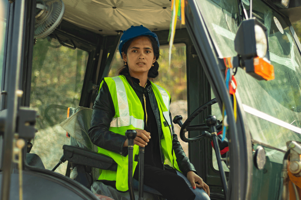 Sushma, a young woman from Nepal, wears a yellow vest and blue helmet as she sits inside the excavator she operates.  