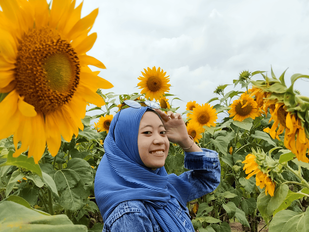 Kumala, a young woman from Indonesia, stands outside in a field of large sunflowers, wearing a blue headscarf and jean jacket, and smiling at the camera. 