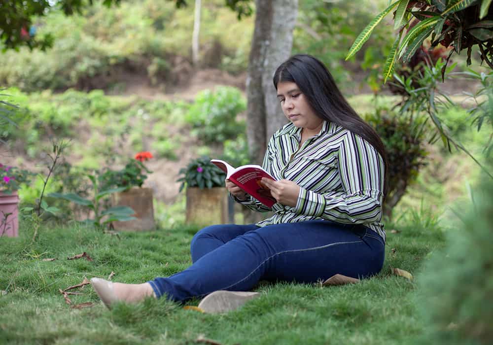Wenddy, wearing jeans and a stripped blouse, sits on the grass and reads a book. 