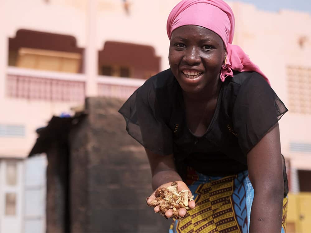 A young African woman smiles at the camera while holding a handful of a light brown substance. She wears a pink headscarf, a black short-sleeved blouse and a blue and yellow patterned skirt. 
