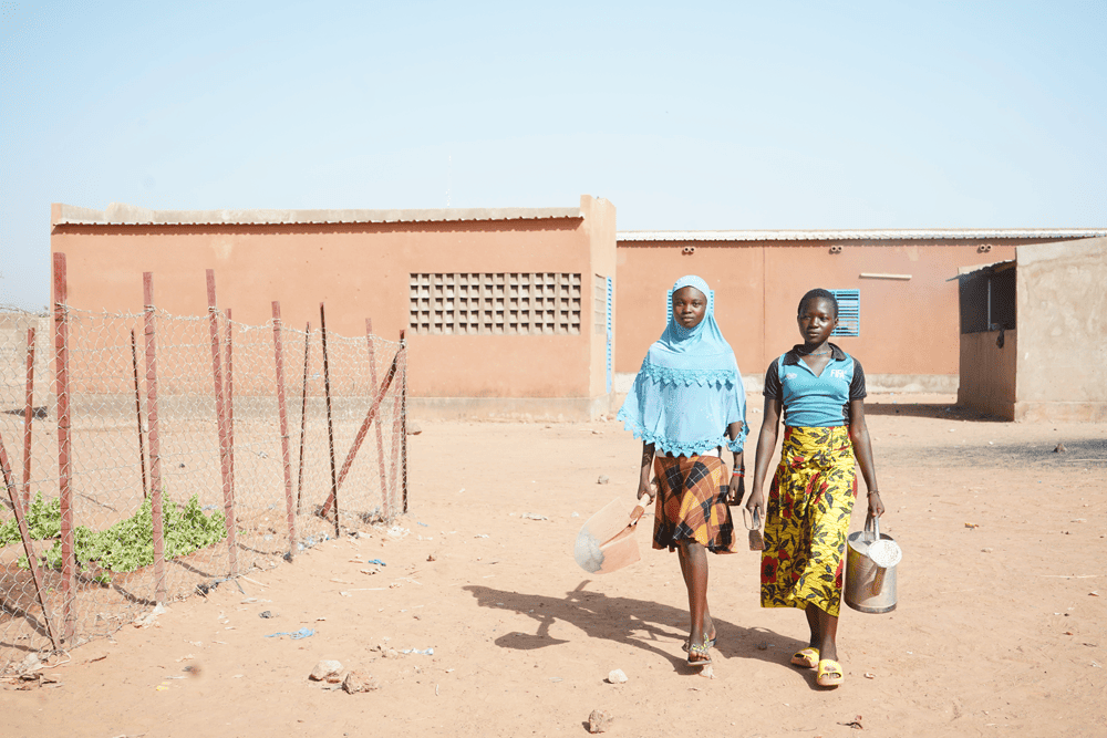 Laeticia and Aminata carry gardening tools on their way to the school garden.
