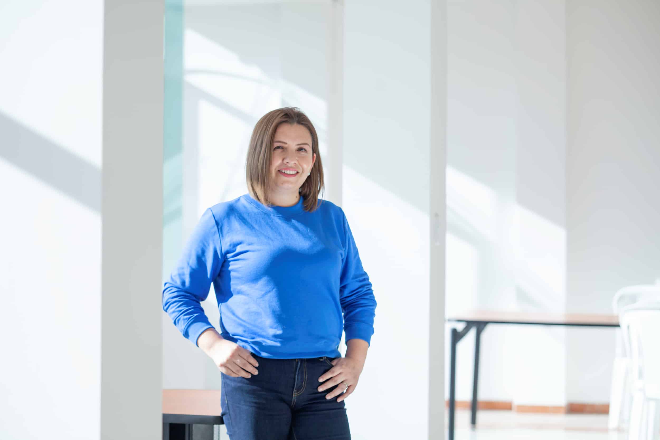 A Latina woman wearing a long-sleeved blue shirt and dark blue jeans stands smiling in front of a white wall. 