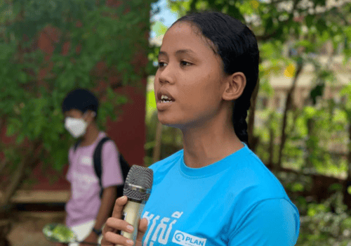 A young Asian woman speaks into a microphone outdoors. She is wearing a blue Plan shirt and standing in front of trees with green leaves.