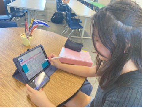 A photo of me, Mona, sitting at a circular school desk while looking at an iPad and an iPhone. 