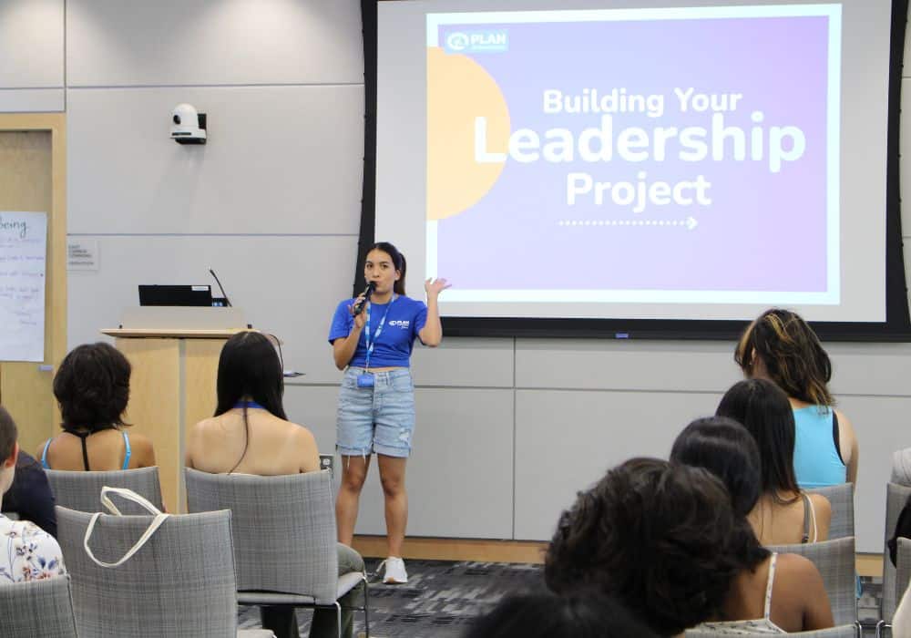 A Plan employee stands in front of a projector that reads “Building Your Leadership Project” and speaks to a group of YLA participants. 