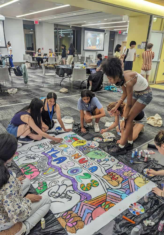 A group of girls sit on an office floor and paint a banner that says, “Plan USA YLA 2023.”